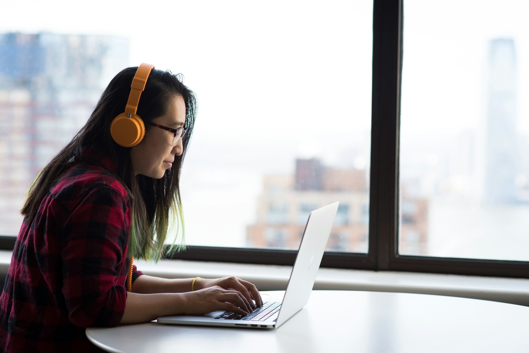 A woman with colorful hair and trendy clothes works from her apartment with high-rise towers faintly visible through her windows