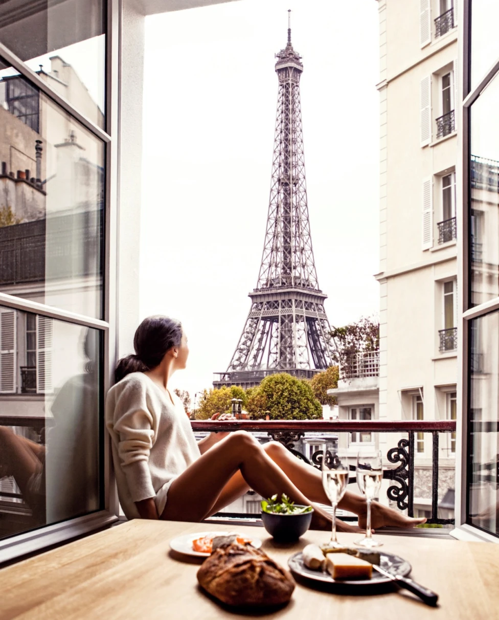 A woman sits along a balcony opening looking up at the Eiffel Tower, a small spread of fine foods and two glasses of white wine sitting on a table just behind her.