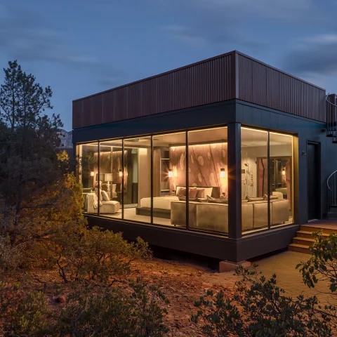 The exterior of a room at Ambiente in the middle of nature at dusk, with floor-to-ceiling windows showcasing a bed and couch inside.