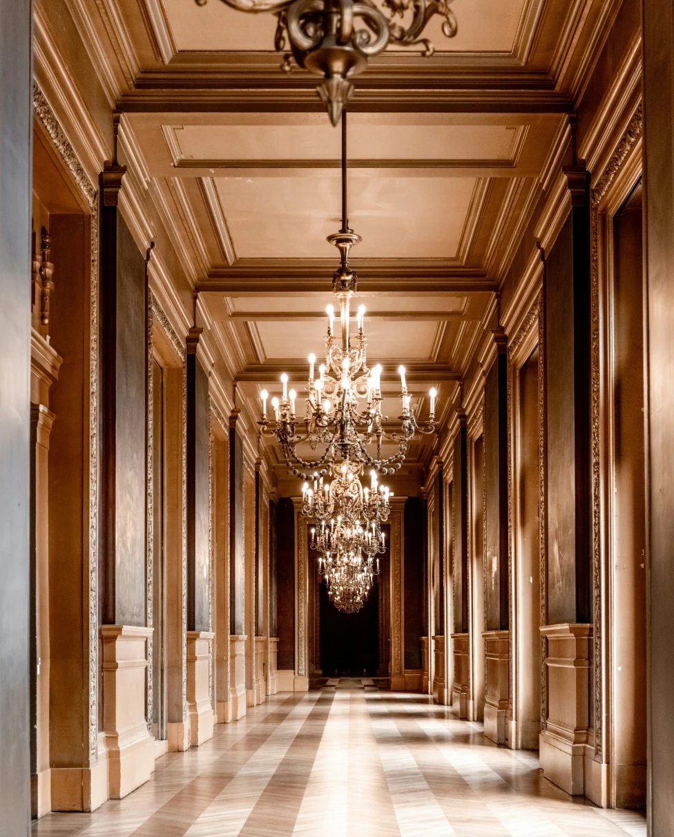 Grand chandeliers hanging from a ceiling in a hallway with striped tile flooring and detailed paneling. 
