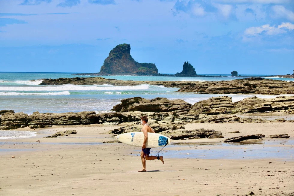 surfer runs down a rocky beach shore towards the water with rock island formations in the distance