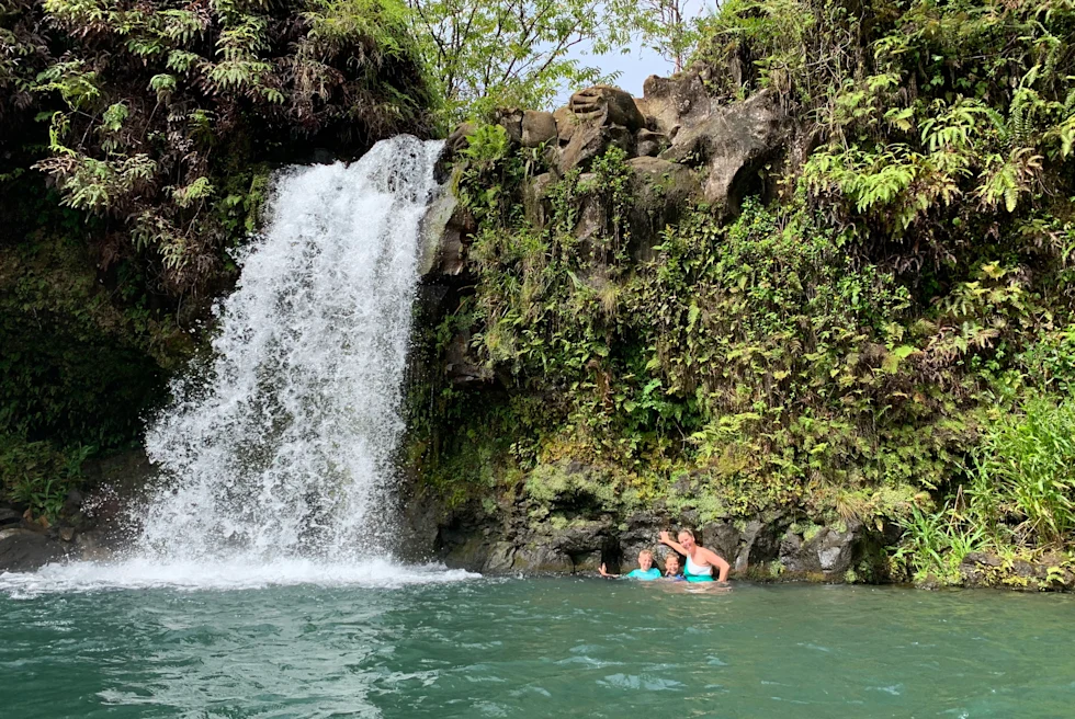 Three people standing in body of water next to a waterfall during daytime