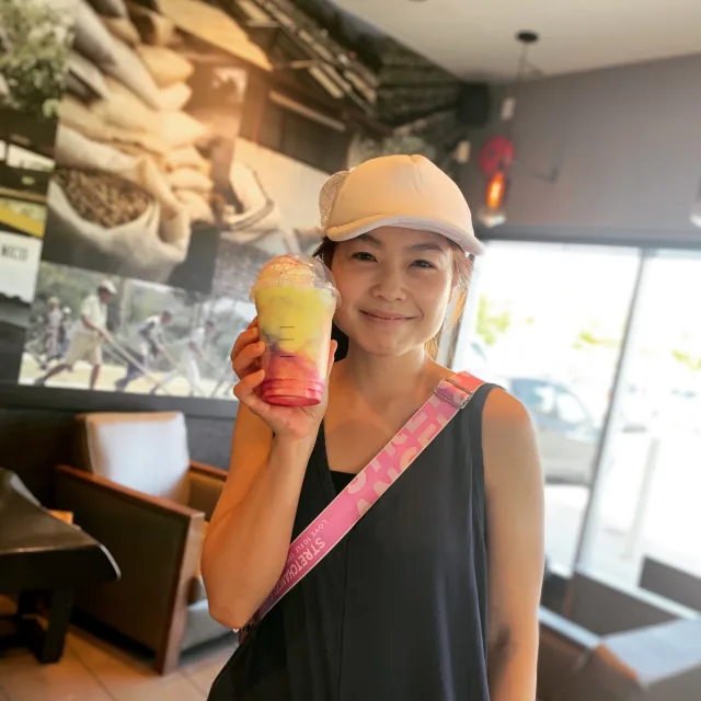 Picture of Daisy wearing a black tank top, white baseball cap and pink crossbody bag with a drink in hand standing inside of a cafe