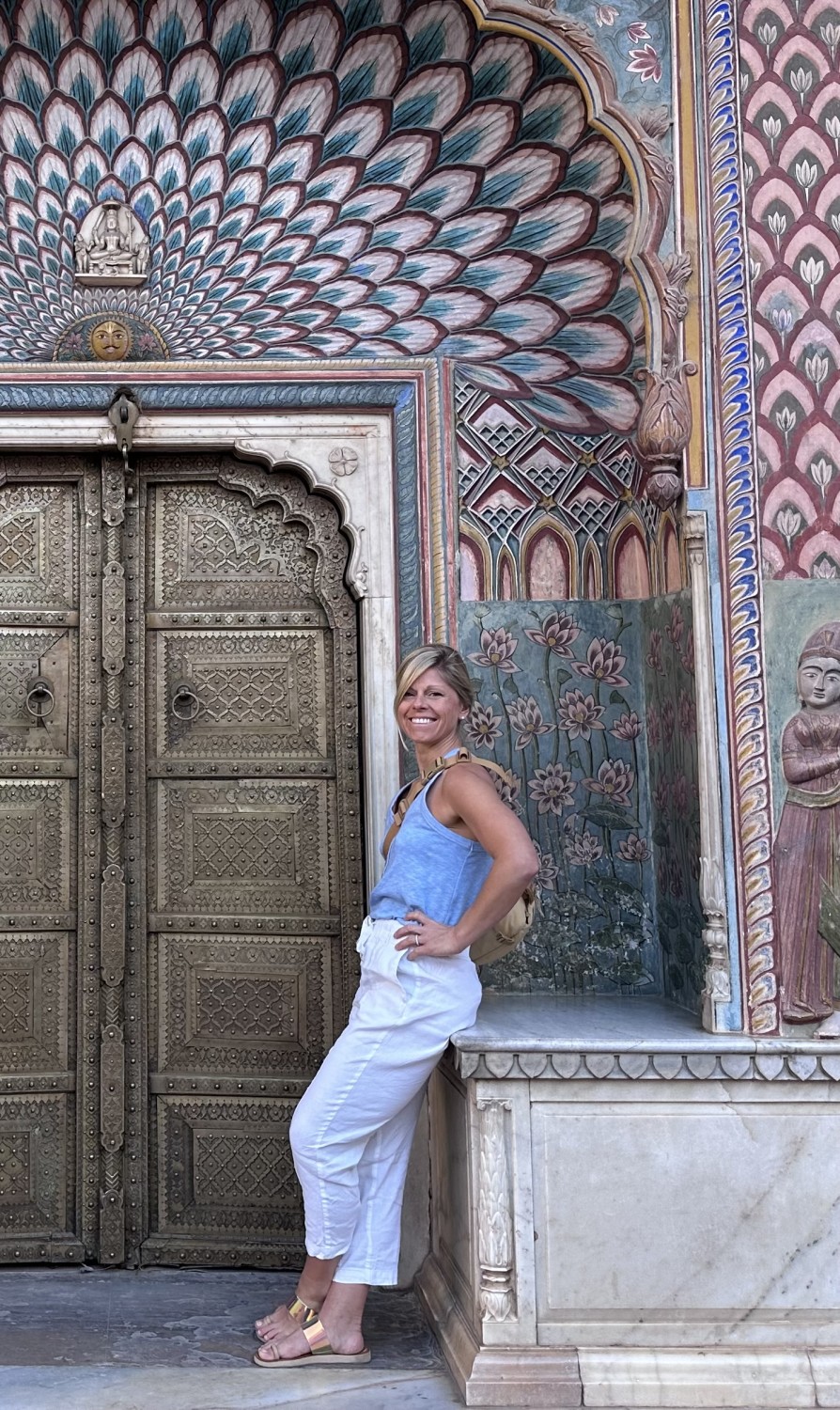 a blonde woman wearing blue pants anf a blue shirt stands in front of an ornate door and a colorful archway
