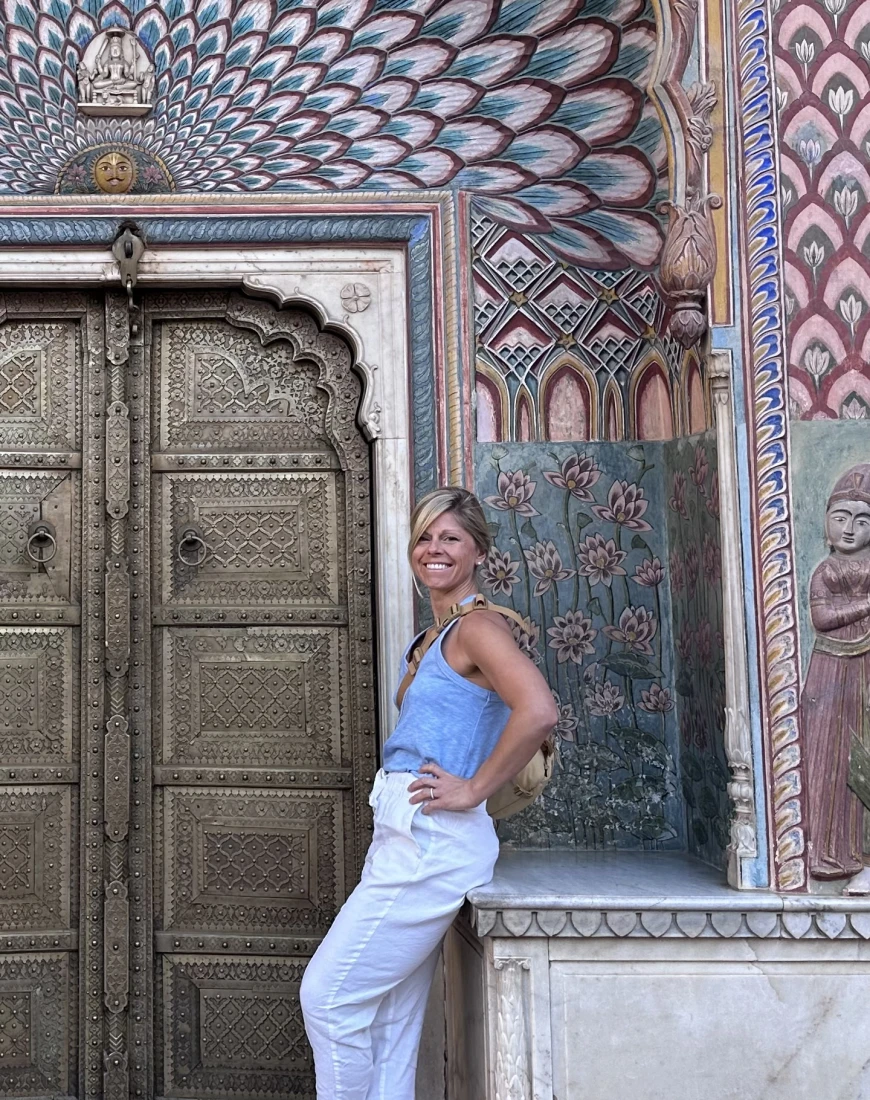 a blonde woman wearing blue pants anf a blue shirt stands in front of an ornate door and a colorful archway