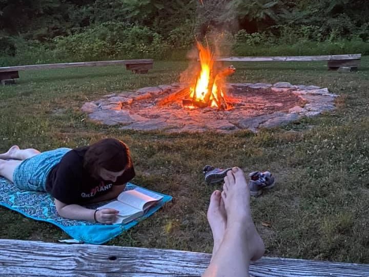 A woman reading a book by camp fire and feet of another one relaxing. 