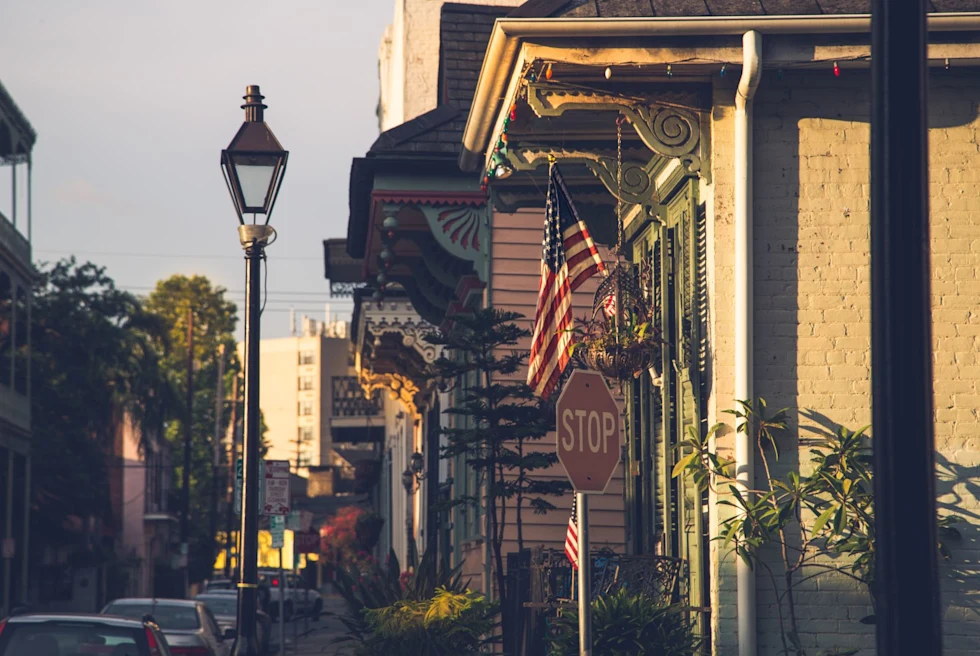 A view of a stop sign, the American flag, a stone building, street lamp and city view in the background.