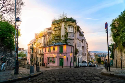 A French street corner near Montmartre Paris hotels with a pink vine-covered building, cobblestone streets, street lamps and a view of the city. 