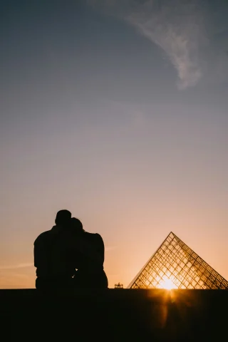 A picture of couple sitting in front of a see-through pyramid watching sunset.