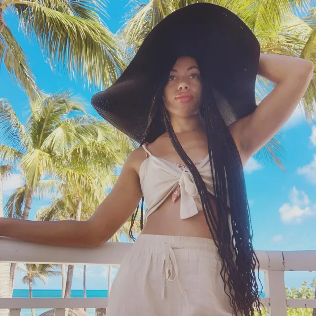 Picture of Maya wearing black sun hat and white outfit posing in front of palm trees on a sunny day