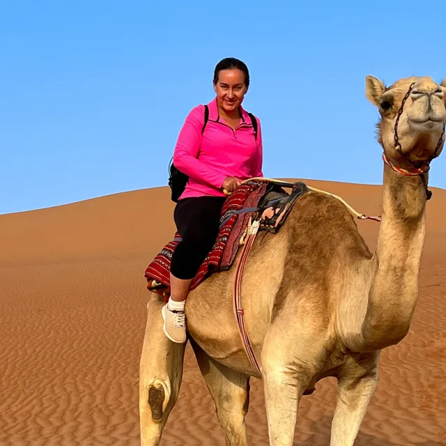 Stephanie wearing a pink top and black pants riding a camel in the desert