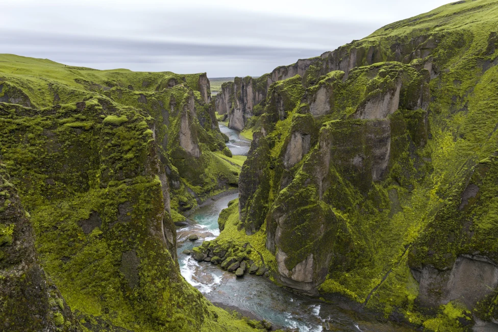 Green moss on tall, large gray rocky mountains with a gushing river flowing through the center on an Iceland itinerary. 