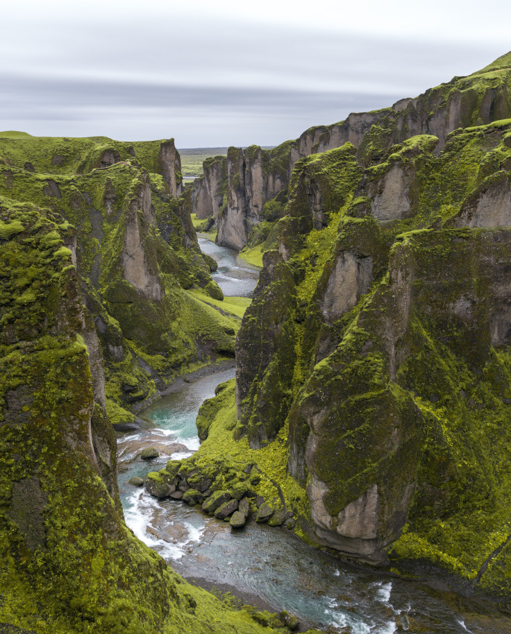 Green moss on tall, large gray rocky mountains with a gushing river flowing through the center on an Iceland itinerary. 