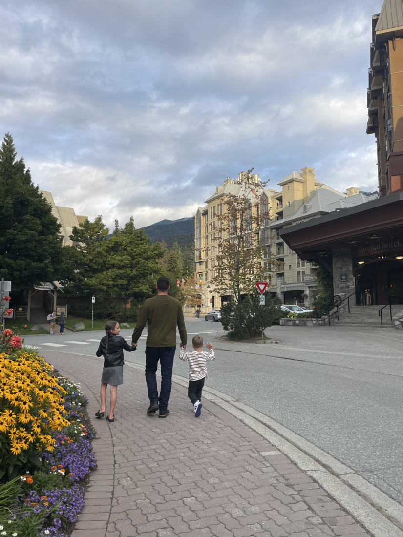 A man and two children walking through Whistler Village near a road, flowers, pine trees and buildings.