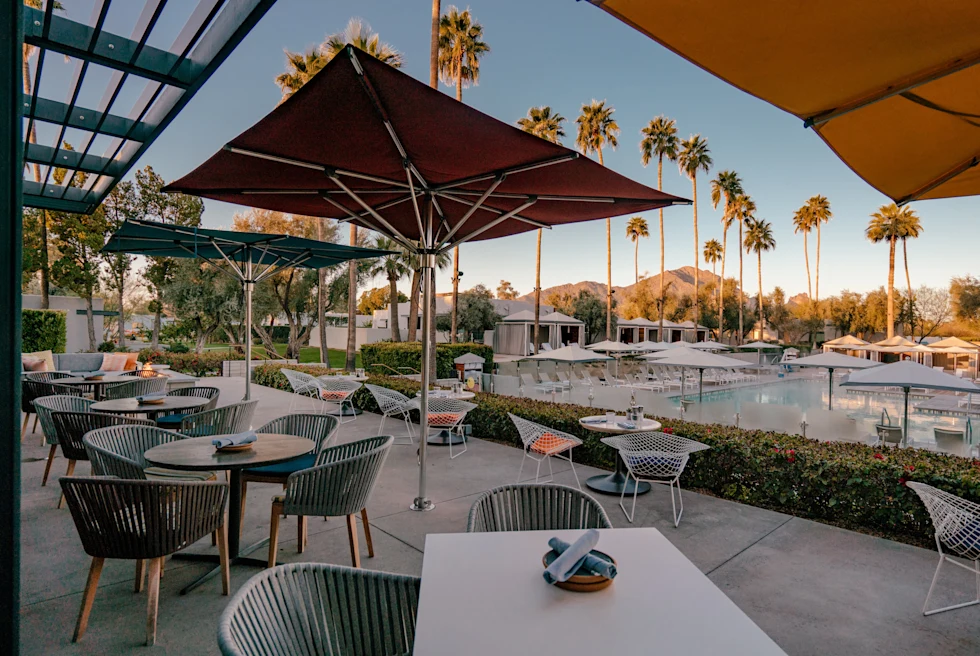 tables and chairs on a patio next to a pool with palm trees