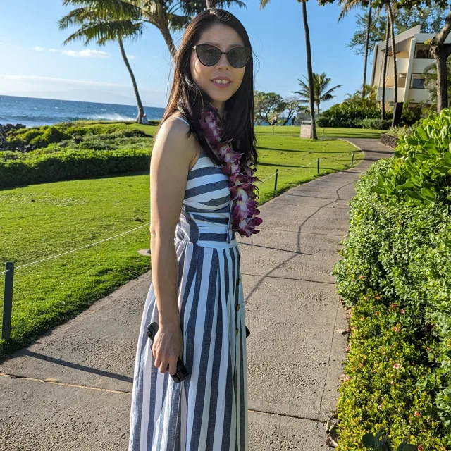 Picture of Alysia in white and blue striped dress on a sidewalk with green grass, palm trees, and the ocean in the background