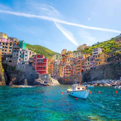 Orange, yellow and blue colorful houses sitting on a rocky cliff overlooking a blue ocean with white boats in Cinque Terre, Italy.