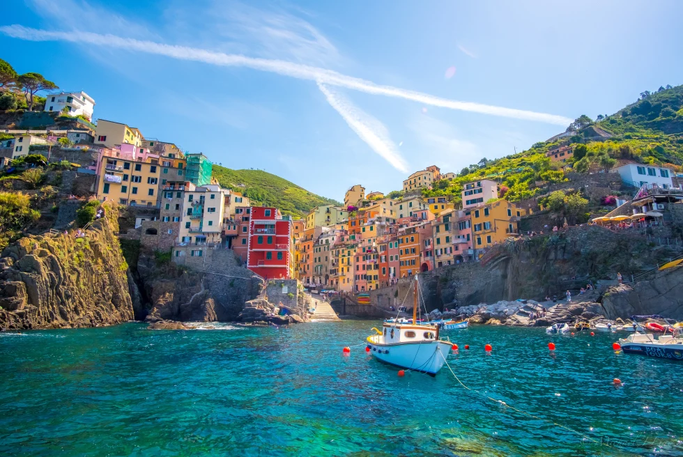 Orange, yellow and blue colorful houses sitting on a rocky cliff overlooking a blue ocean with white boats in Cinque Terre, Italy.