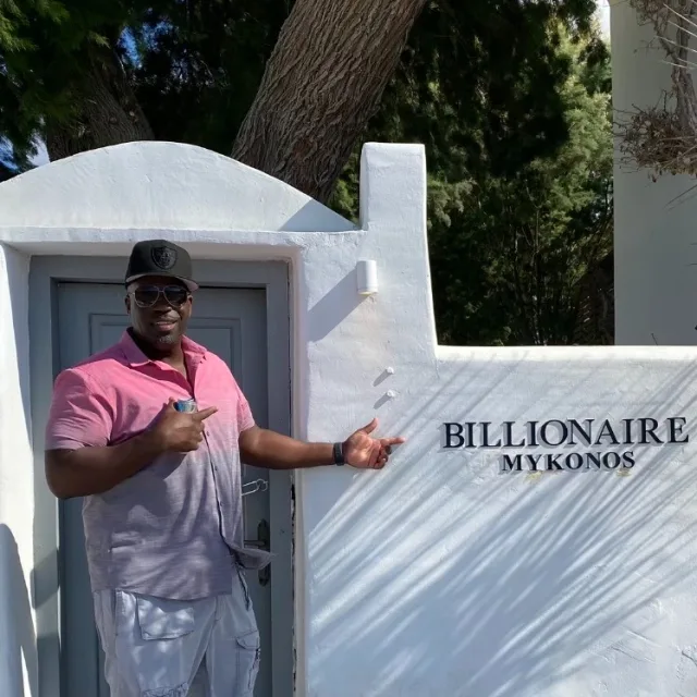 Travel advisor Darnell Hill pointing towards house's name plate in writing BILLIONAIRE MY KONOS