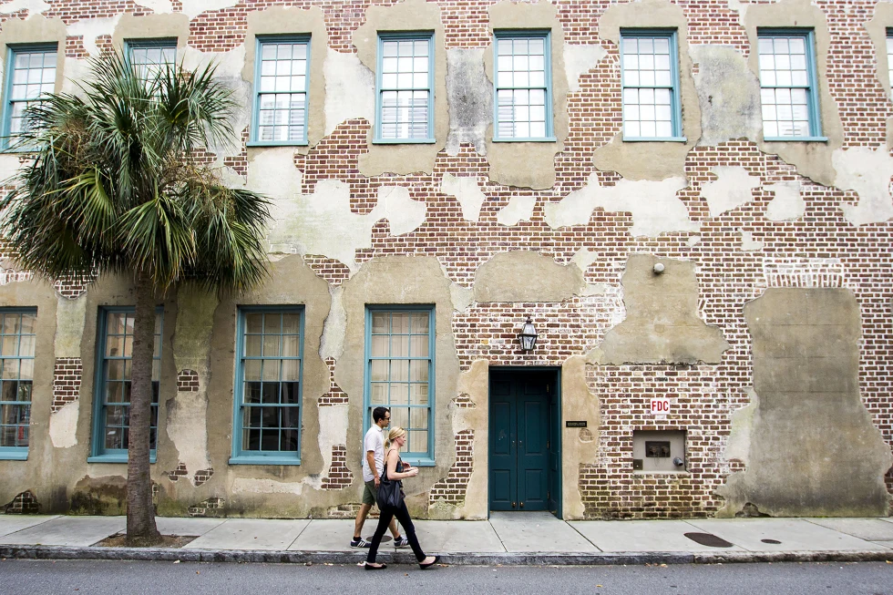 two people walking in front of brick wall next to a palm tree