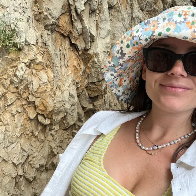 Travel Advisor Maggie Reynolds with a floral hat, green bikini top and white shirt in front of a rock.