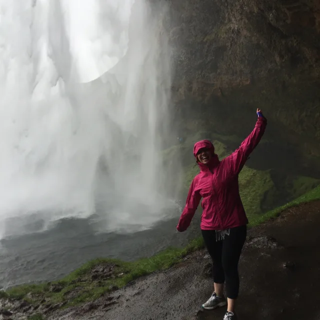 Becky wearing a pink raincoat standing next to a giant waterfall in mountains