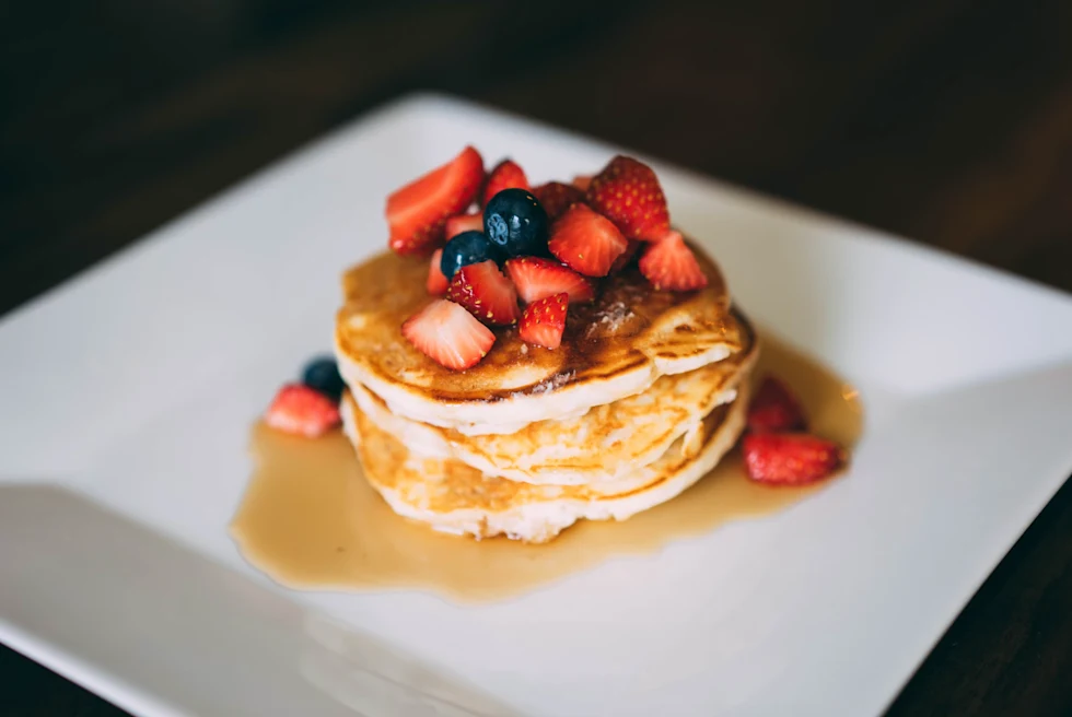 A stack of pancakes covered in maple syrup and berries on a white square plate.
