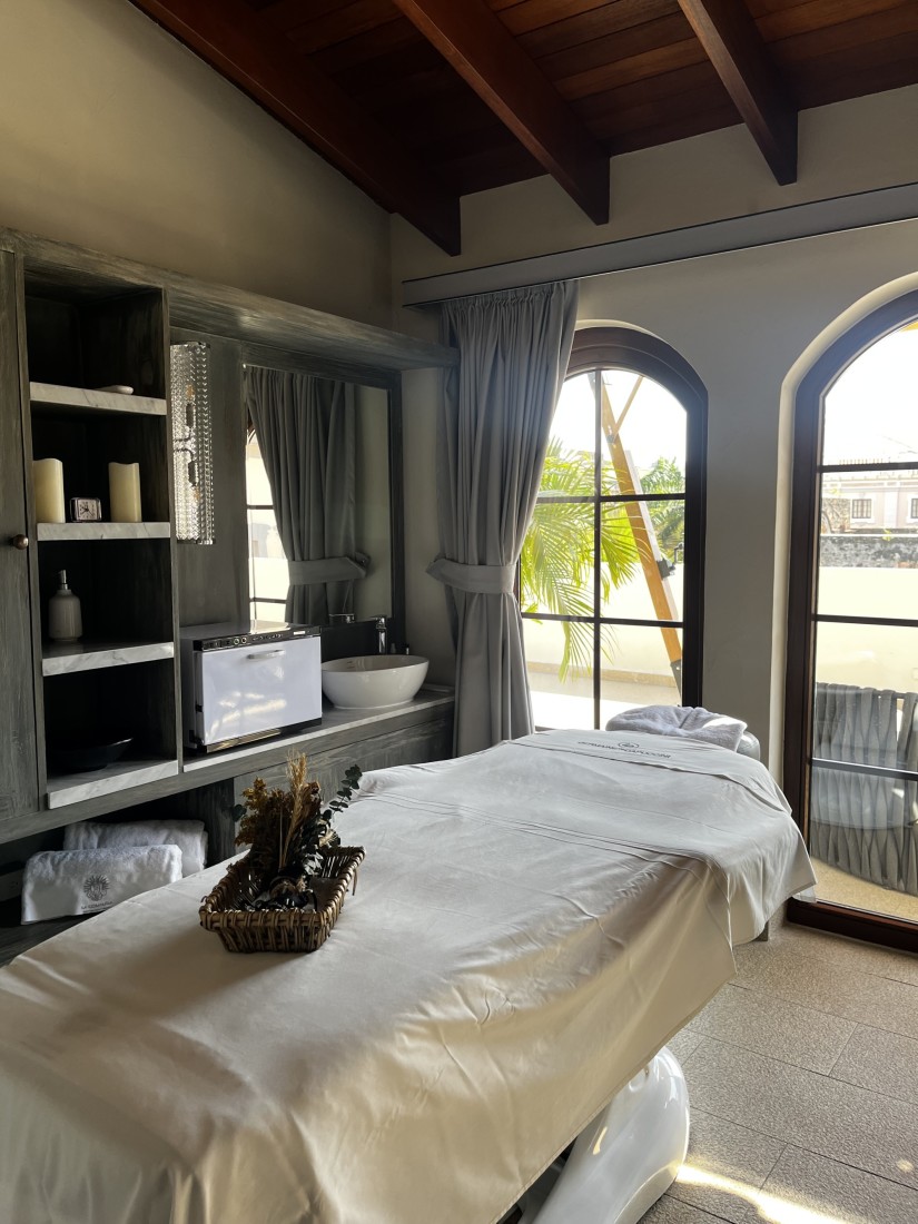 A massage table with shelving, a mirror with a sink and arched windows with a curtain beneath a wood paneled ceiling. 