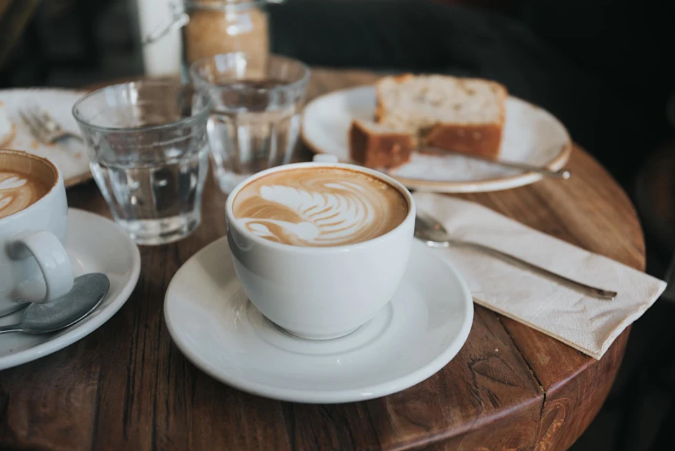 a latte with milk froth art and a pastry on a wood table with other glasses and cutlery