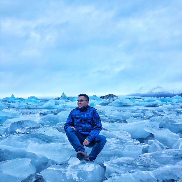 Man wearing blue jacket and jeans sitting on ice with a cloudy sky