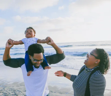 Man holding a baby on his shoulders on a beach with a woman, all three laughing.