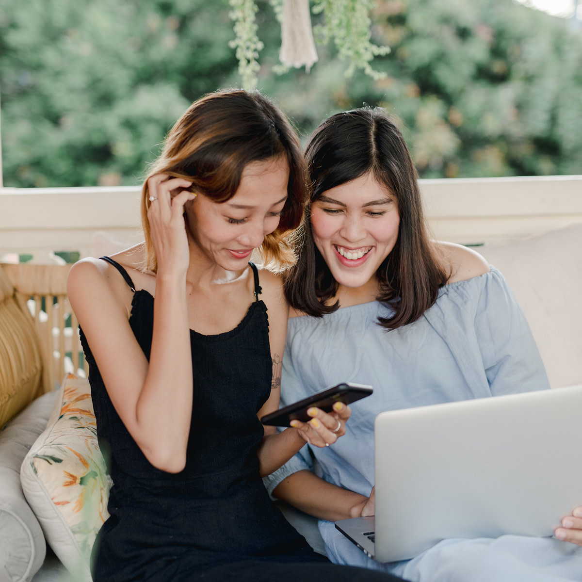 two women sitting next to each other and smiling at their computer and phone with a green leafy background