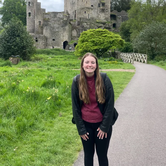 Allison wearing a red top, black shirt and black pants in front of a castle and green grass. 