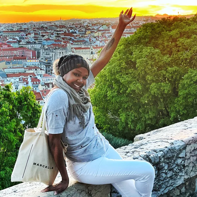 Travel Advisor Rashida Joy Allen sitting on a stone wall in white pants and a grey shirt with buildings and trees in the background at sunset.