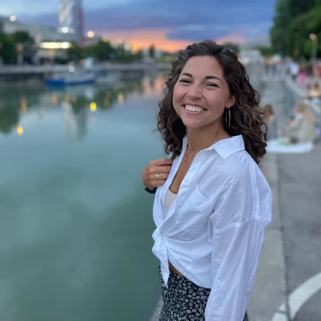 Emily wearing a white top and smiling in front of a river with a cityscape and sunset in the distance.