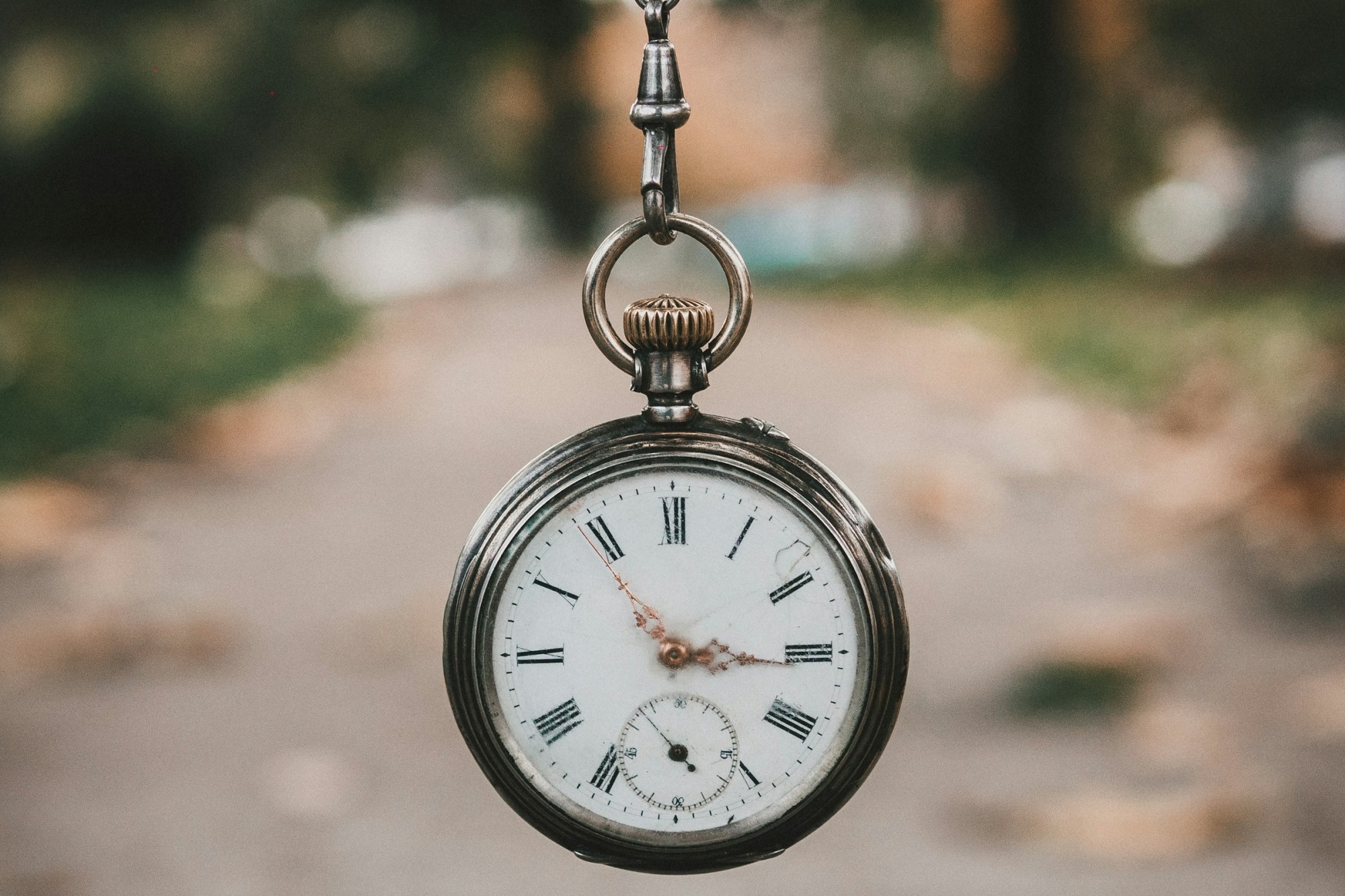 A classic timepiece is held at the end of a chain in focus while a path with leaves stretches out ahead, likely at a university
