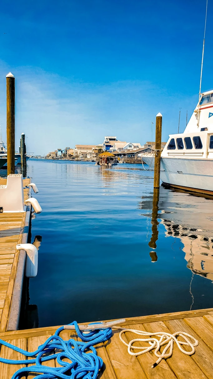 The image depicts a tranquil dockside setting with clear skies and calm waters, featuring a wooden dock and a white boat moored to the side.