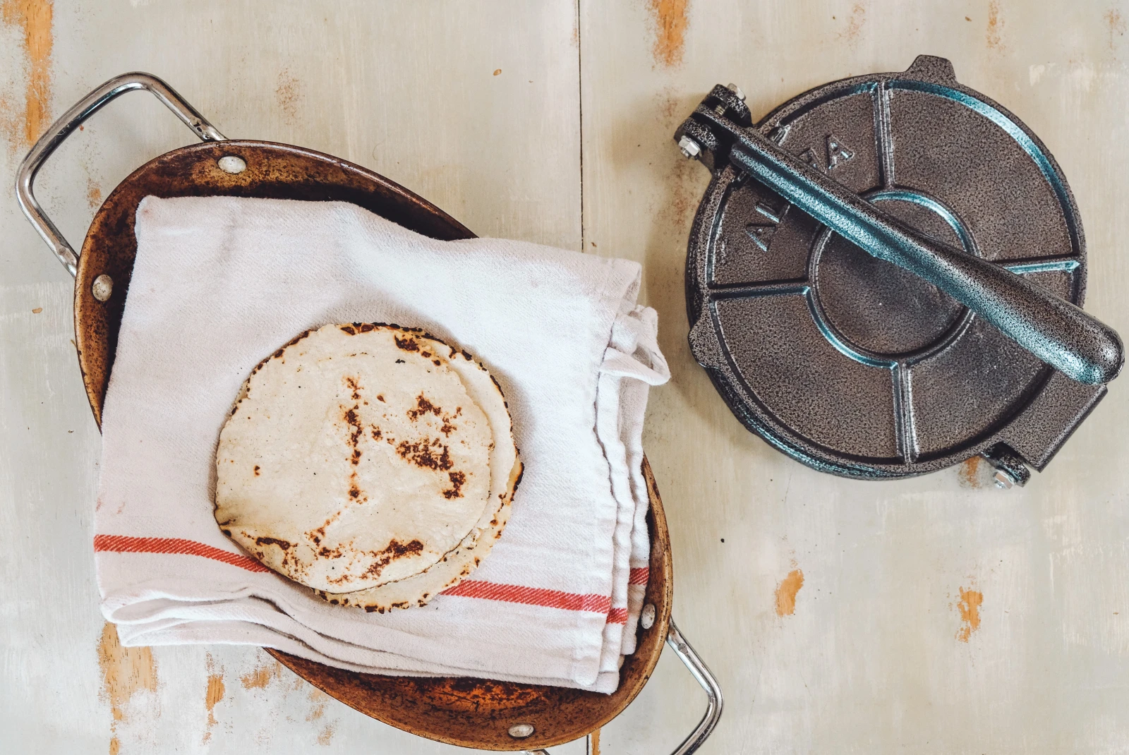 Tortillas on a white kitchen towel in a pan next to a cast iron tortilla press