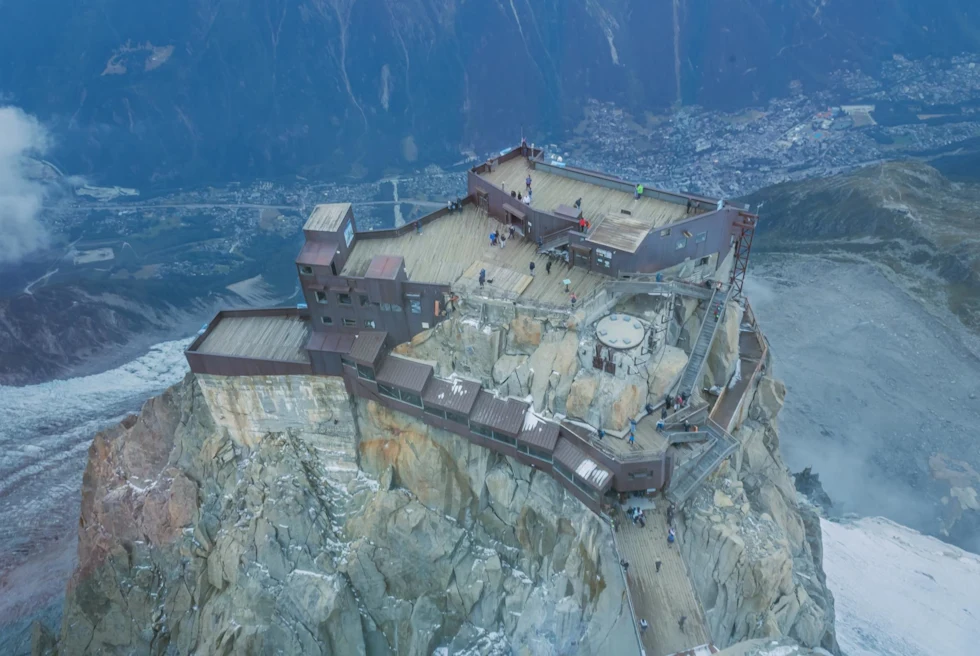 a wooden deck with people a top of towering cliff in a snowy mountain range