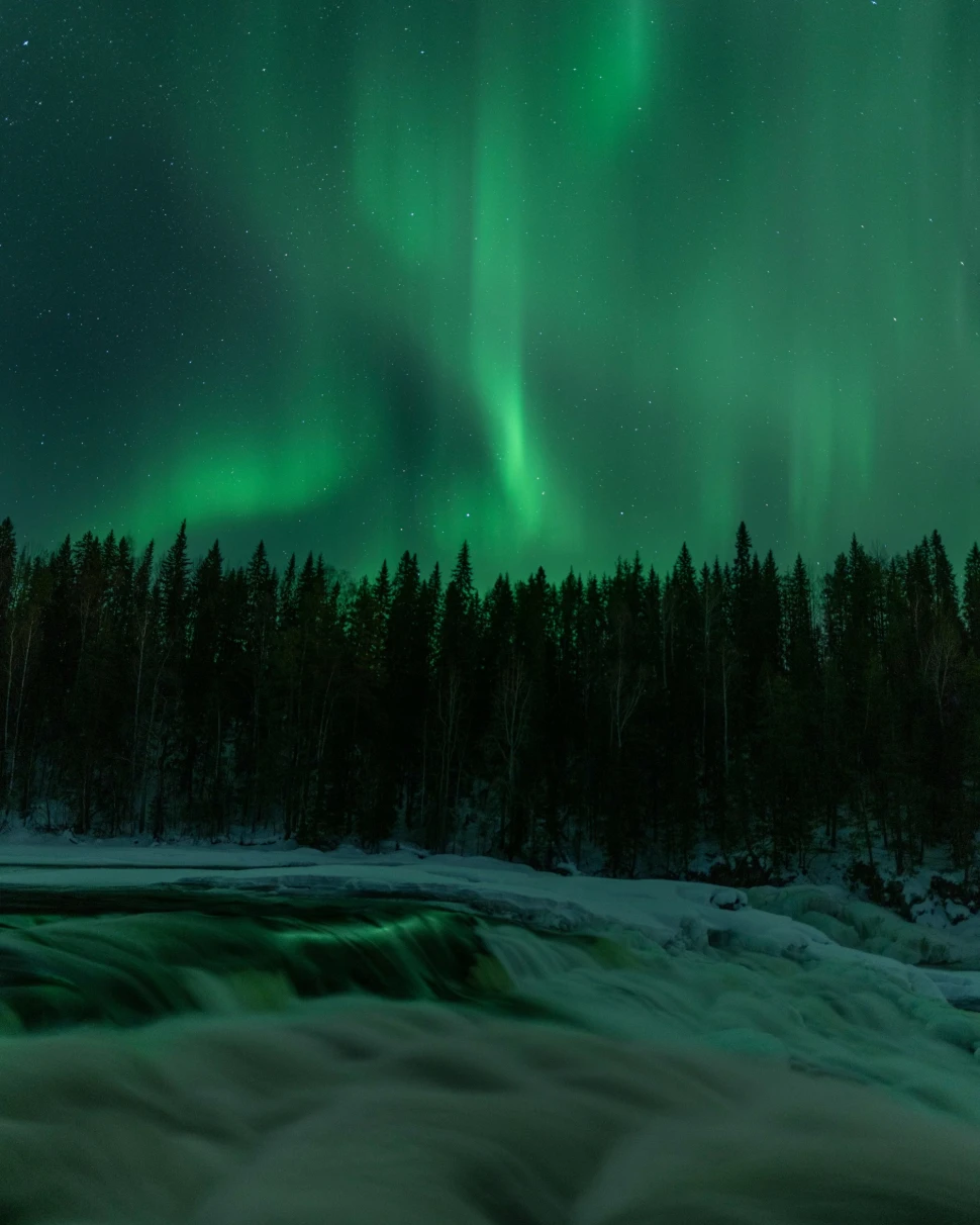 The image captures the stunning, green northern lights over a snowy landscape with a river.