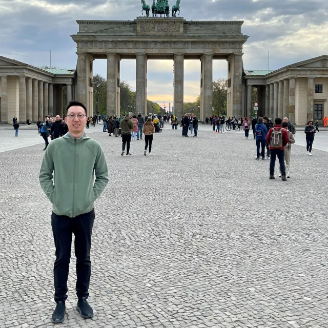 Picture of James wearing a green top and black pants in front of the Brandenburg Gate with tourists in the background.