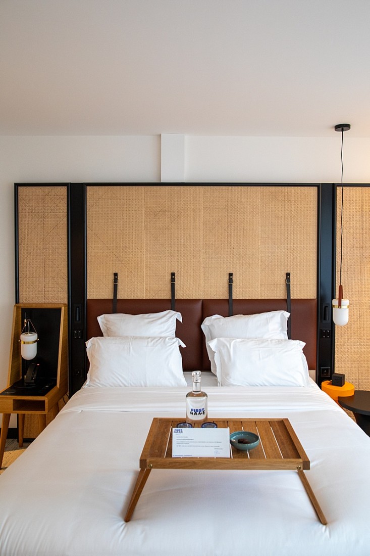 A hotel suite bedroom with a white bed, brown leather headboard, side tables, pendant lighting and a wooden tray placed on top of the bed with a card and water carafe. 