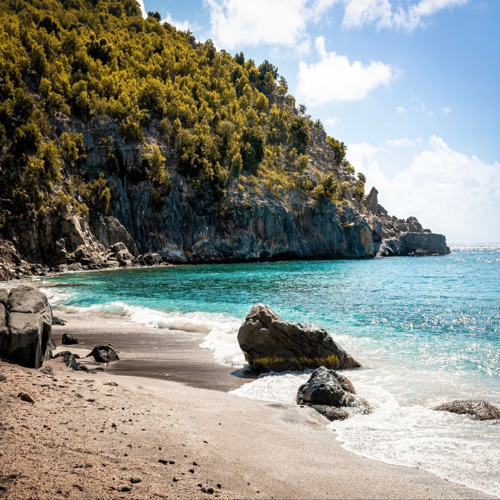 A tranquil beach with turquoise waters, green hillside and rocky formations under a sky with scattered clouds.