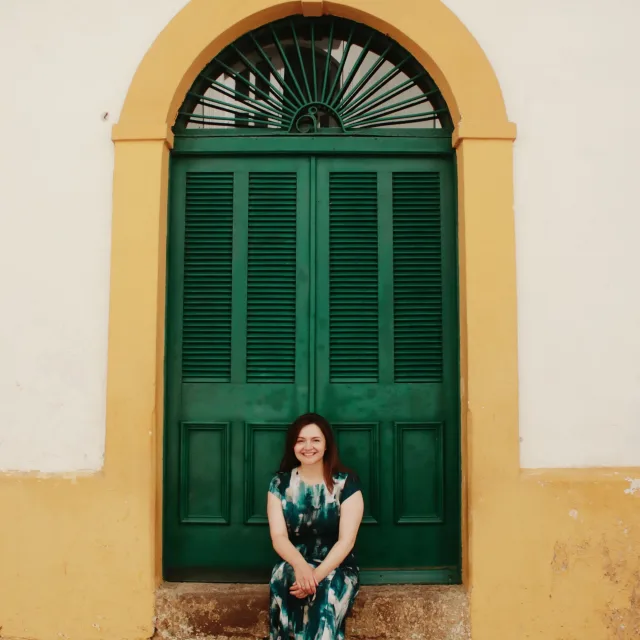 Travel advisor Pam Hughet sits on the stoop of a large green door with yellow boarder wearing a green and white dress