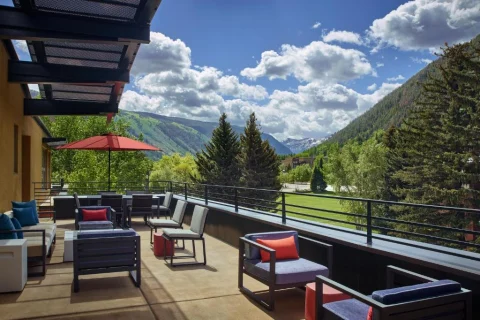 This image depicts a hotel terrace at The Limelight Aspen with various options for lounge seating and umbrellas. There is a stunning view of tree covered mountains and a cloudy blue sky in the background. 