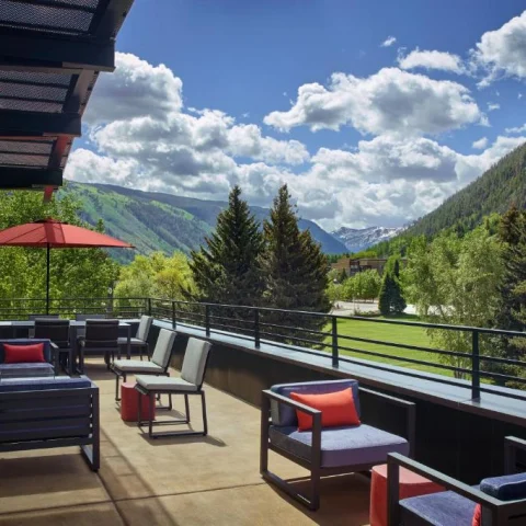 This image depicts a hotel terrace at The Limelight Aspen with various options for lounge seating and umbrellas. There is a stunning view of tree covered mountains and a cloudy blue sky in the background. 