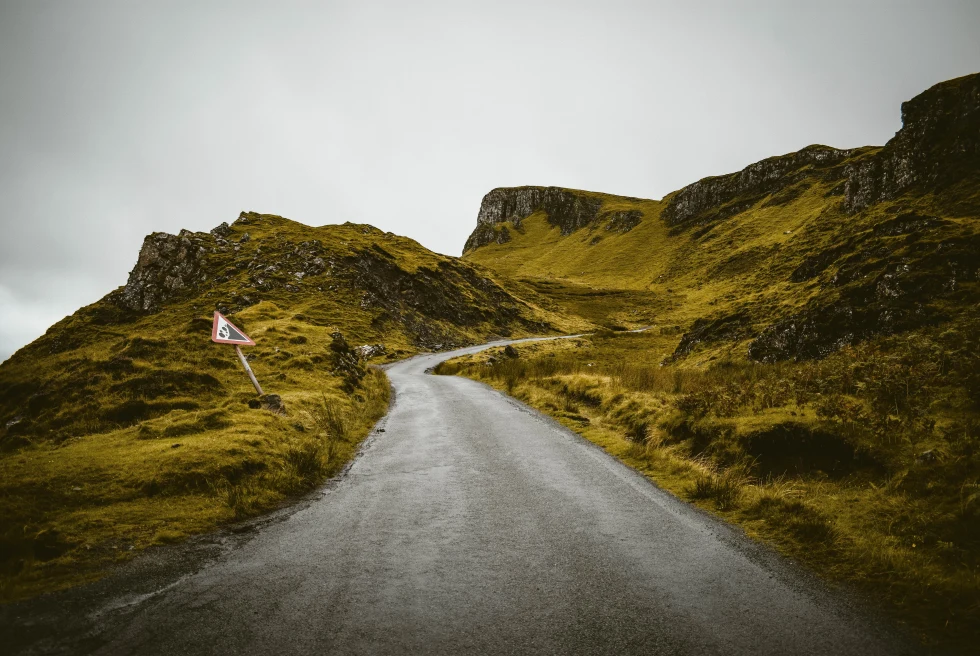 long windy black road going uphill with green and brown rocky rolling hills