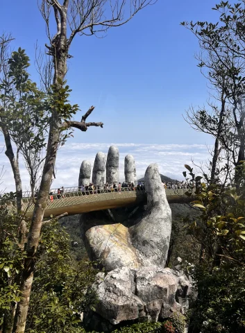 A view of a large stone hand with a bridge built onto it and people walking across. There are also two sets of tall trees and a forest in the surrounding areas. 