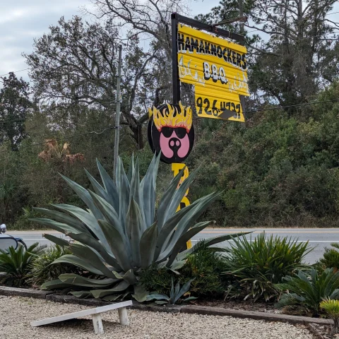 A yellow sign for a restaurant surrounded by trees and aloe vera plants on the side of the road outside. 