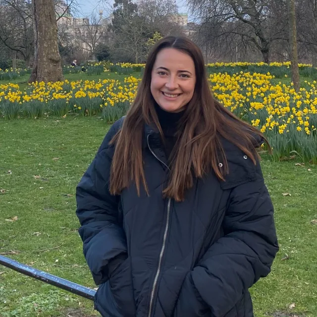 Travel Advisor Julia DeJesus smiles wearing a black coat in a park with a yellow flower bed